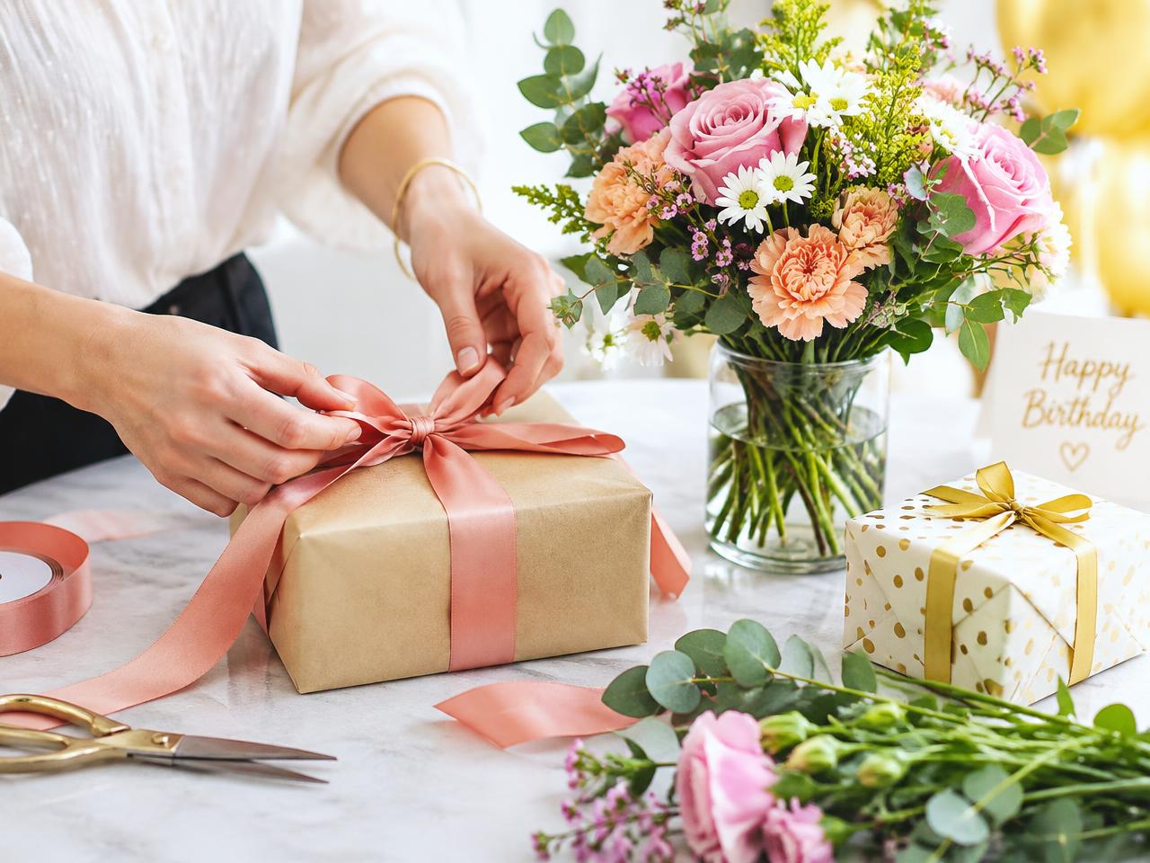 Hands tying a ribbon on a gift beside a bouquet of pink flowers